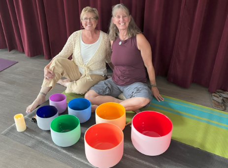 Two women sitting on the floor in front of colorful sound bowls, smiling at the camera